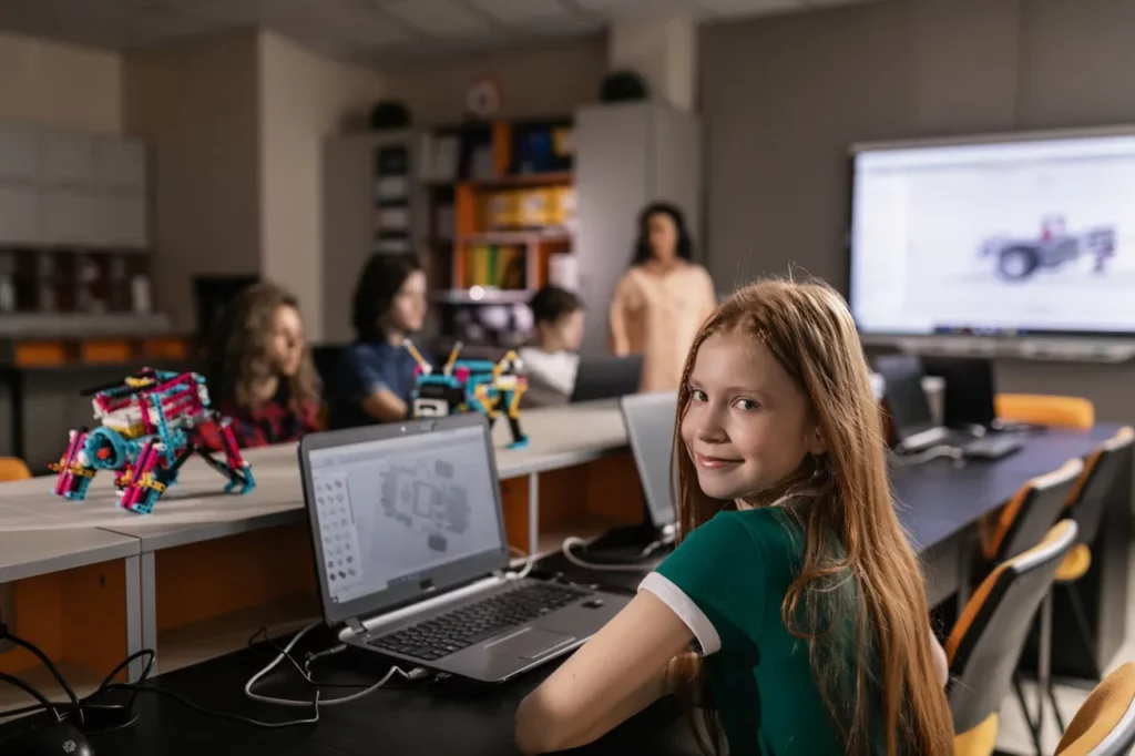 Girl in foreground sitting at laptop with robots and other students in the background during an engineering robotics event